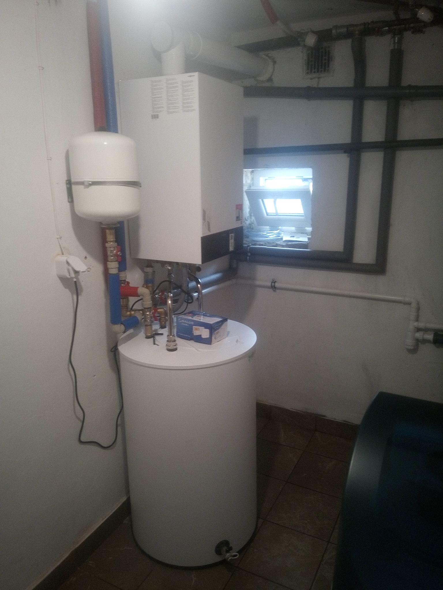 Biały piec gazowy z zasobnikiem, rurami i armaturą w kotłowni. Widoczne okno dachowe i instalacje. Ujęcie z dołu, podkreślające złożoność systemu.