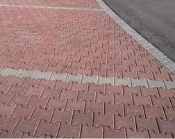 Czerwona nawierzchnia ułożona z kostki brukowej o charakterystycznym wzorze, oddzielona jasnoszarą linią od asfaltowej drogi. Widoczne łączenia kostek i faktura powierzchni.