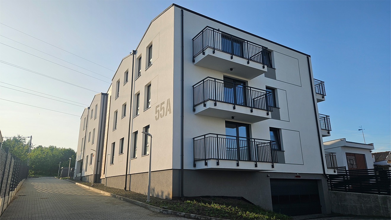Nowoczesny, trzypiętrowy budynek mieszkalny z balkonami o czarnych barierkach, oświetlony porannym słońcem. Widoczny numer budynku 55A i brama garażowa.