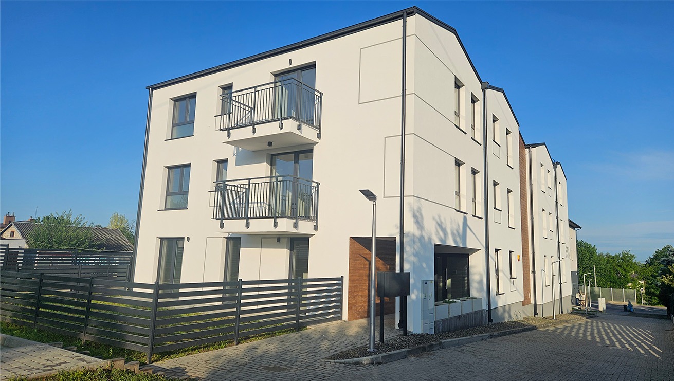 Nowoczesny, trzypiętrowy budynek mieszkalny z balkonami i jasną elewacją, widziany z poziomu chodnika. Uwagę zwraca minimalistyczny design i staranne wykończenie.