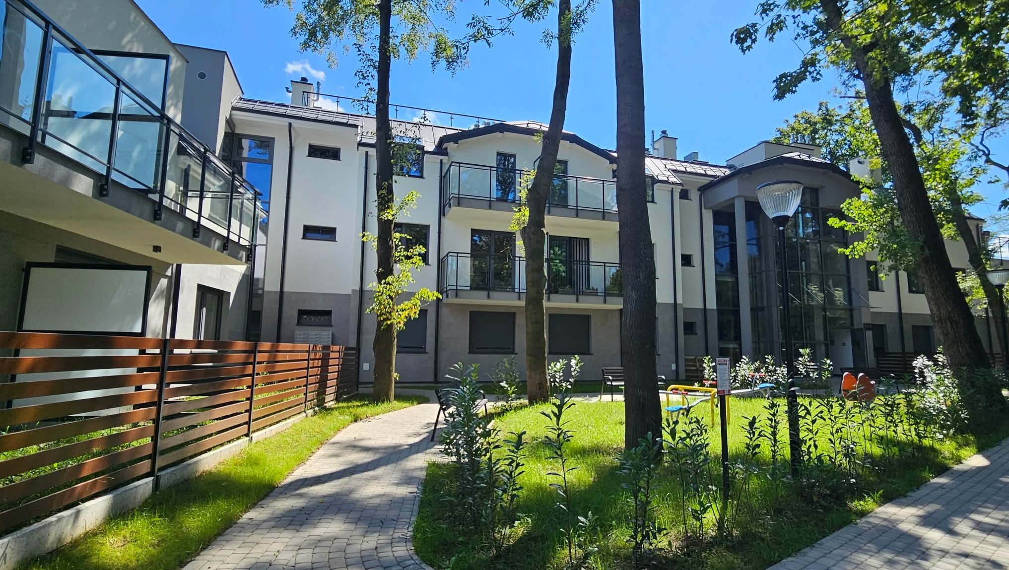 Nowoczesny, trzypiętrowy budynek mieszkalny z balkonami i szklanymi balustradami, otoczony zielenią i ścieżką z kostki brukowej w słoneczny dzień.