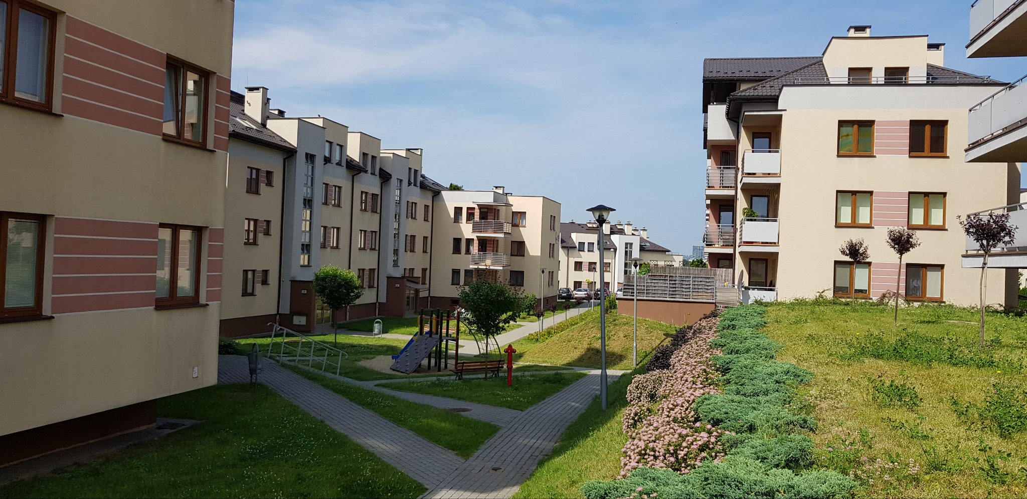 Osiedle budynków wielorodzinnych z placem zabaw i zielenią. Jasna elewacja z brązowymi oknami. Widok na nowoczesną architekturę w słoneczny dzień.