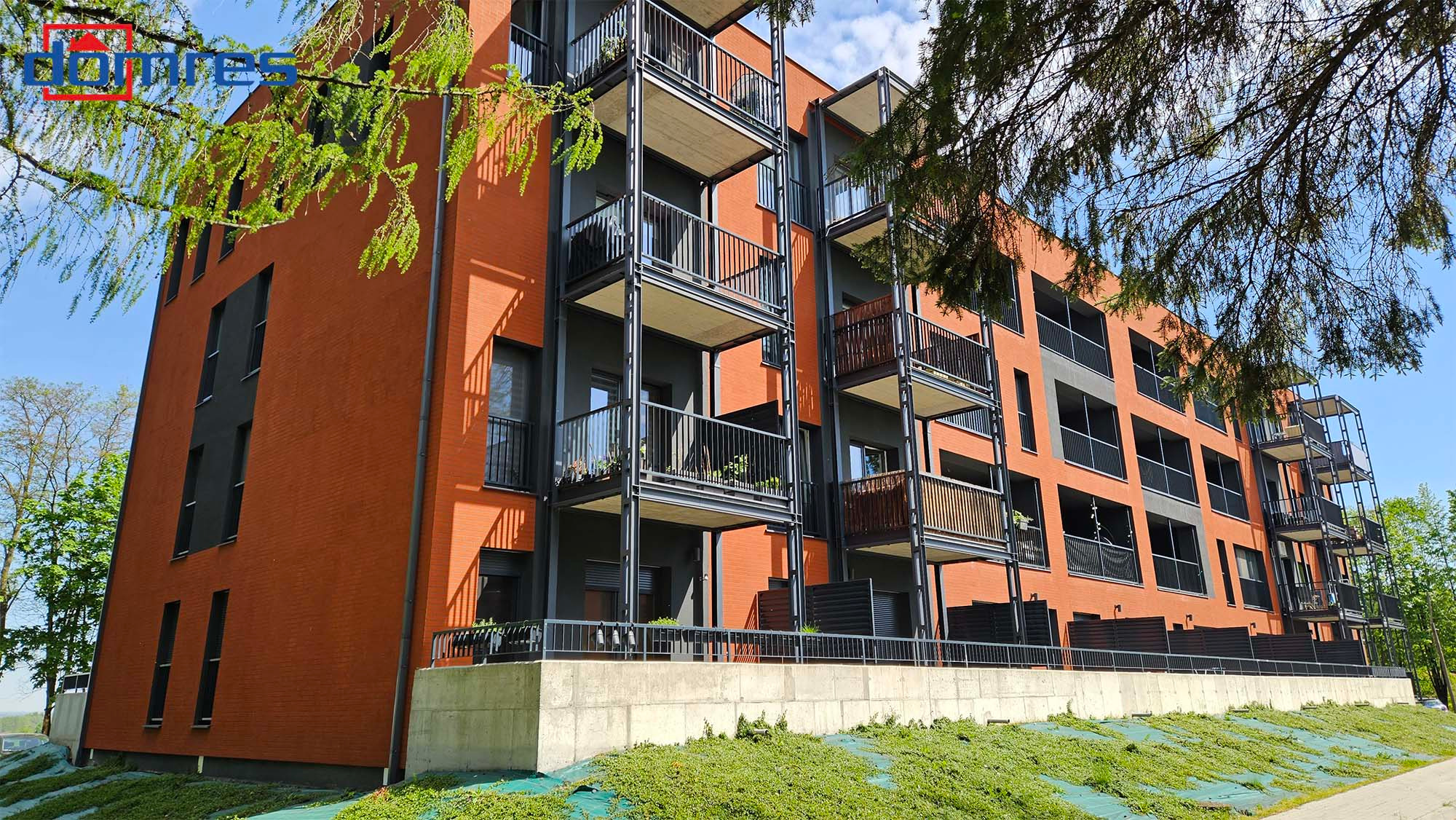 Nowoczesny budynek mieszkalny z ceglaną elewacją i balkonami o stalowej konstrukcji, widok z dołu, zieleń w tle, słoneczny dzień.