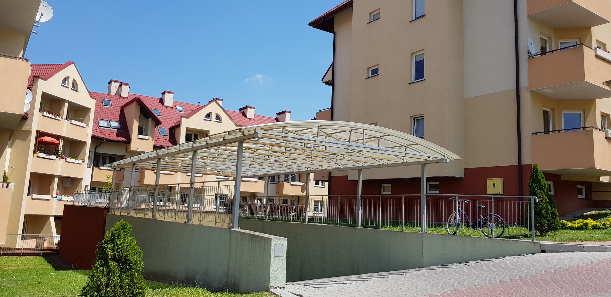 Nowoczesne osiedle z podziemnym parkingiem, zadaszone wejście, jasna elewacja budynków, czerwone dachy, błękitne niebo. Dwa rowery zaparkowane przy barierce.