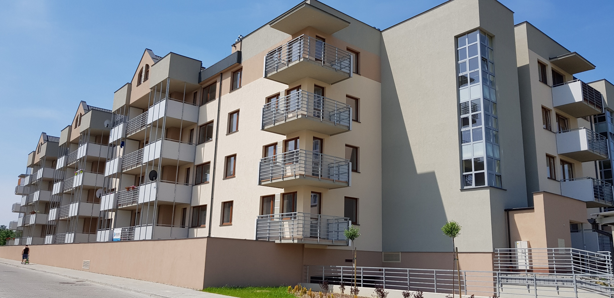 Nowoczesny budynek wielorodzinny z balkonami o metalowych barierkach, jasna elewacja z akcentami w odcieniach beżu i szarości, błękitne niebo w tle.