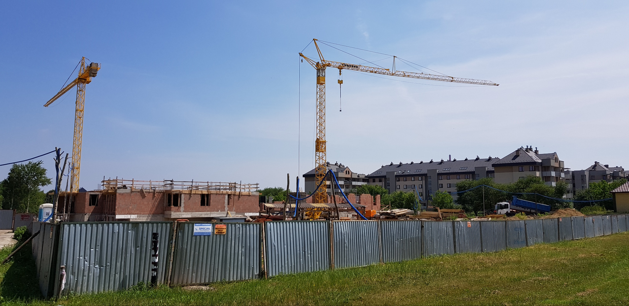 Budowa domu z cegły: widoczne mury, drewniane szalunki, dwa żółte żurawie na tle błękitnego nieba i zabudowy wielorodzinnej. Teren budowy ogrodzony blachą falistą.