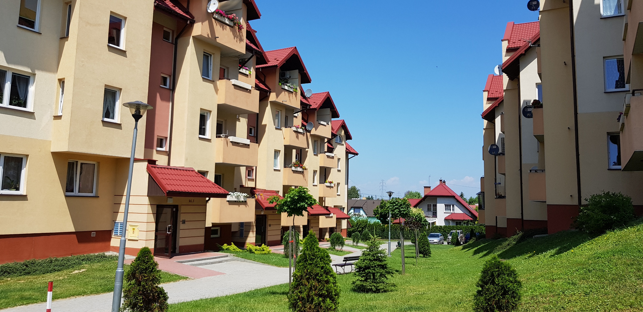 Nowoczesne, beżowe bloki z czerwonymi dachami i balkonami, otoczone zielenią i drzewami w słoneczny dzień. Widoczne detale architektoniczne i zadbany teren.