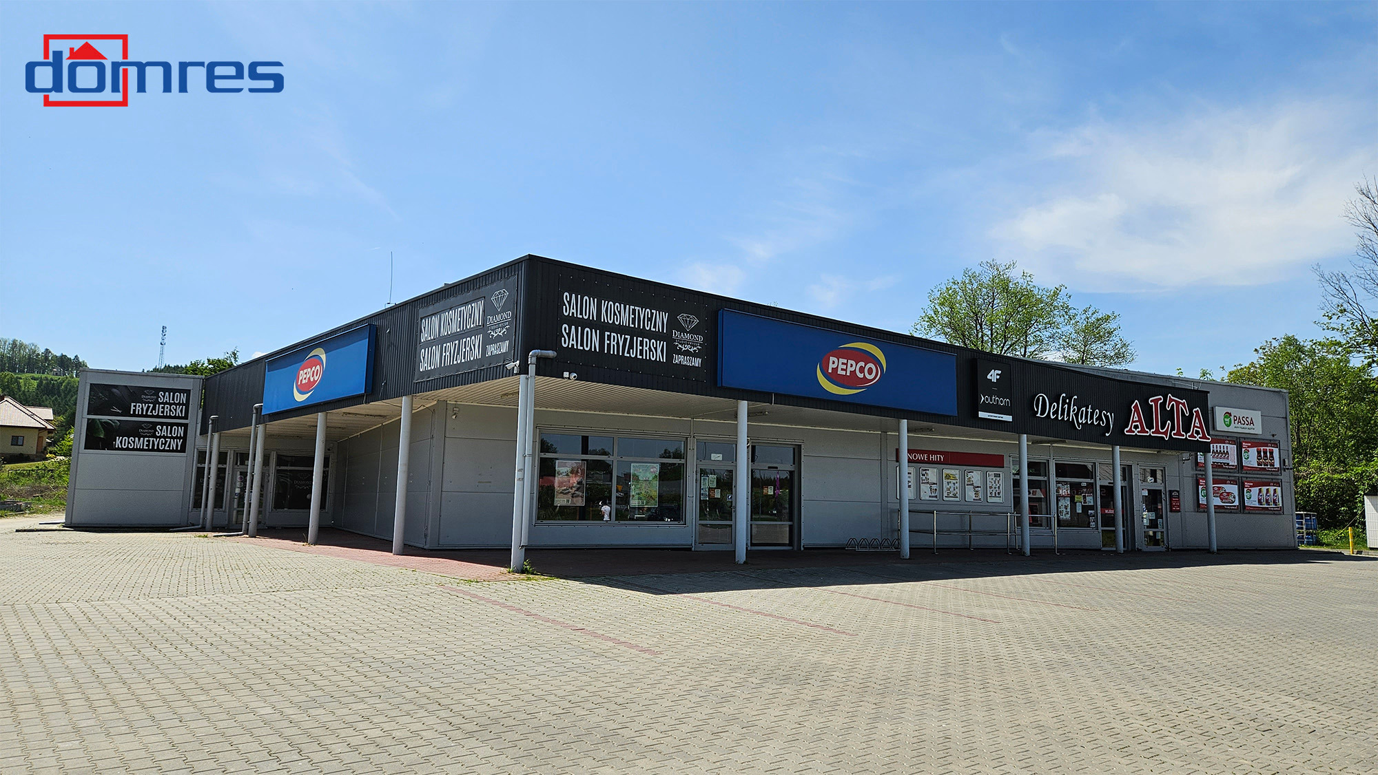 Budynek handlowo-usługowy z salonami kosmetycznymi, fryzjerskimi, sklepem Pepco i delikatesami Alta. Widok z zewnątrz, kostka brukowa na parkingu.
