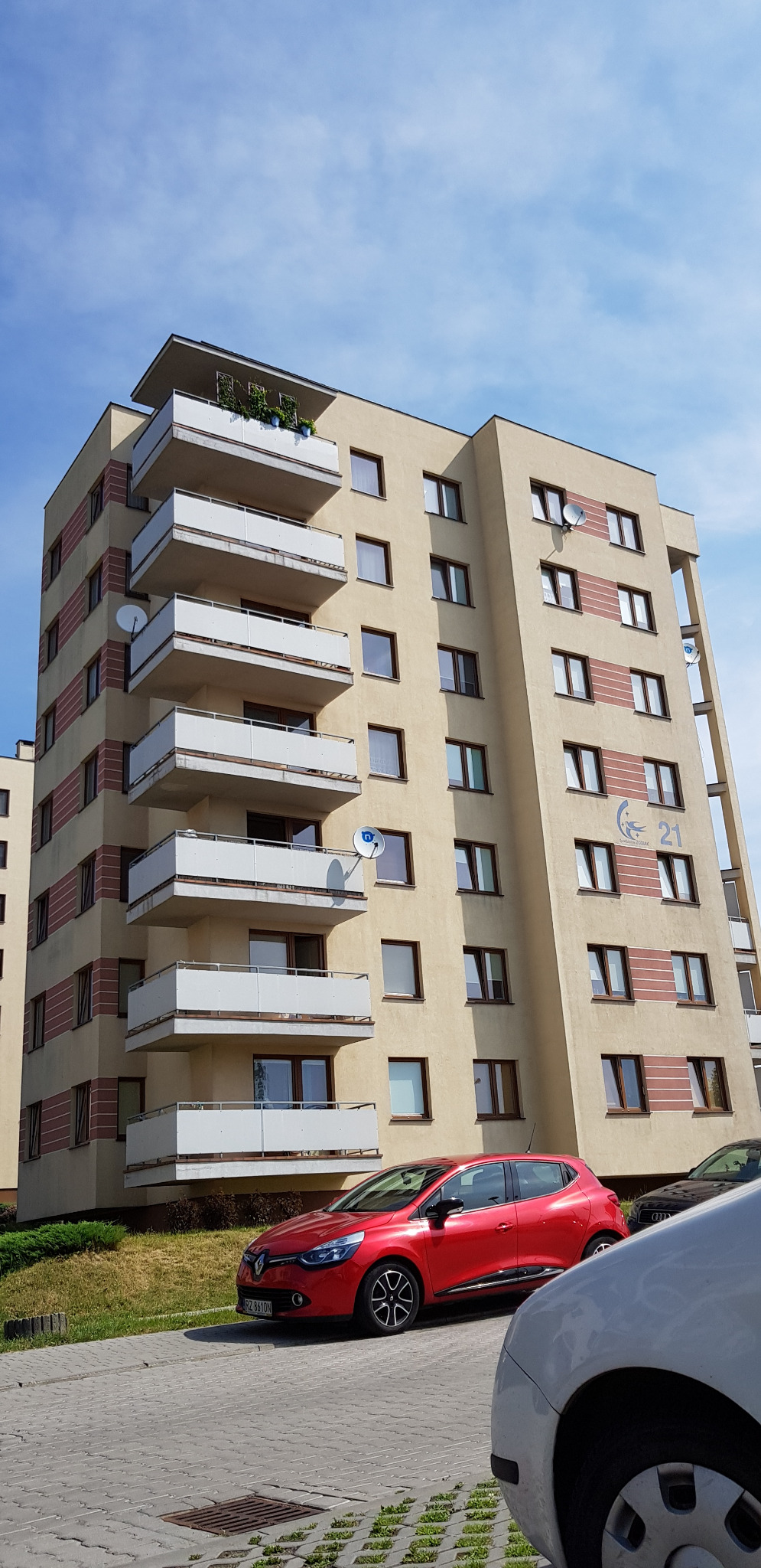 Wielopiętrowy budynek mieszkalny z balkonami, ocieplony w odcieniach beżu i brązu, z widocznymi antenami satelitarnymi i czerwonym samochodem na pierwszym planie.