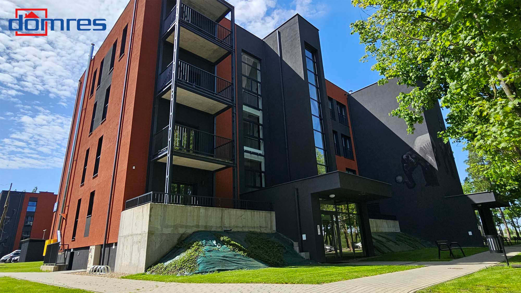 Nowoczesny budynek wielorodzinny z balkonami, wykończony w kolorach cegły i grafitu, z muralem na ścianie. Widok z poziomu chodnika, słoneczny dzień.