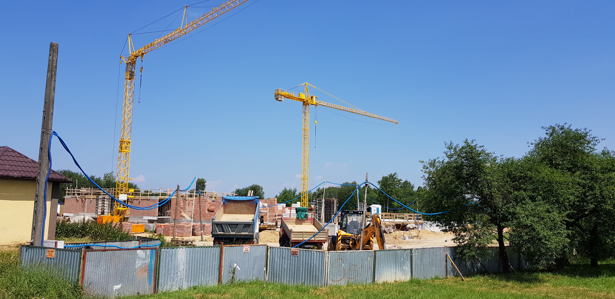Budowa domu jednorodzinnego w toku: widoczne fundamenty, ściany z cegły, dwa żółte dźwigi, ciężarówki z piaskiem i wykopana ziemia. Całość otoczona metalowym płotem.