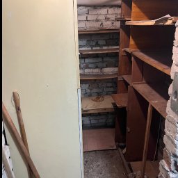 Pusto pl - Zdemontowane, drewniane regały w piwnicy z ceglanymi ścianami. Obok stoi lodówka i leżą drewniane kije. Widoczny gruz i resztki po demontażu.