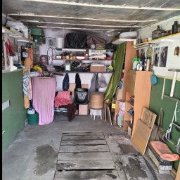 Pusto pl - Zagracony garaż z półkami pełnymi przedmiotów, podłogą z drewnianych desek i zielonymi ścianami. Widoczne narzędzia, meble i różne przedmioty codziennego użytku.