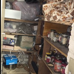 Pusto pl - Zastawiona półka w pomieszczeniu z różnymi przedmiotami, w tym słoikami, pudełkami i workami. Widoczne elementy muru i drewniane półki. Nieporządek.