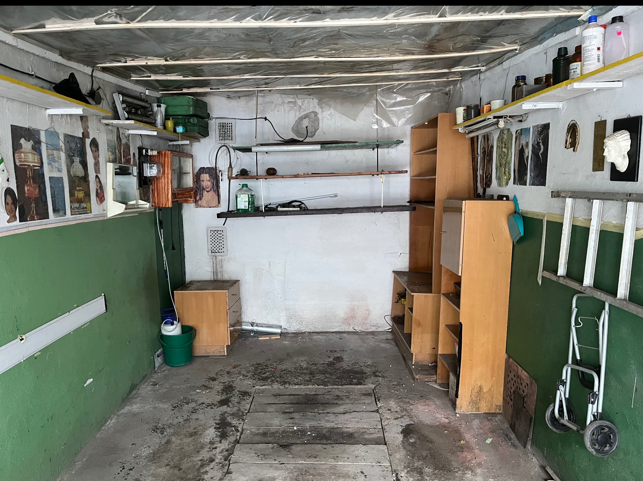 Zagracony garaż z drewnianą podłogą, regałami, drabiną i różnymi przedmiotami na półkach. Ściany pomalowane na zielono i biało, sufit przykryty folią.