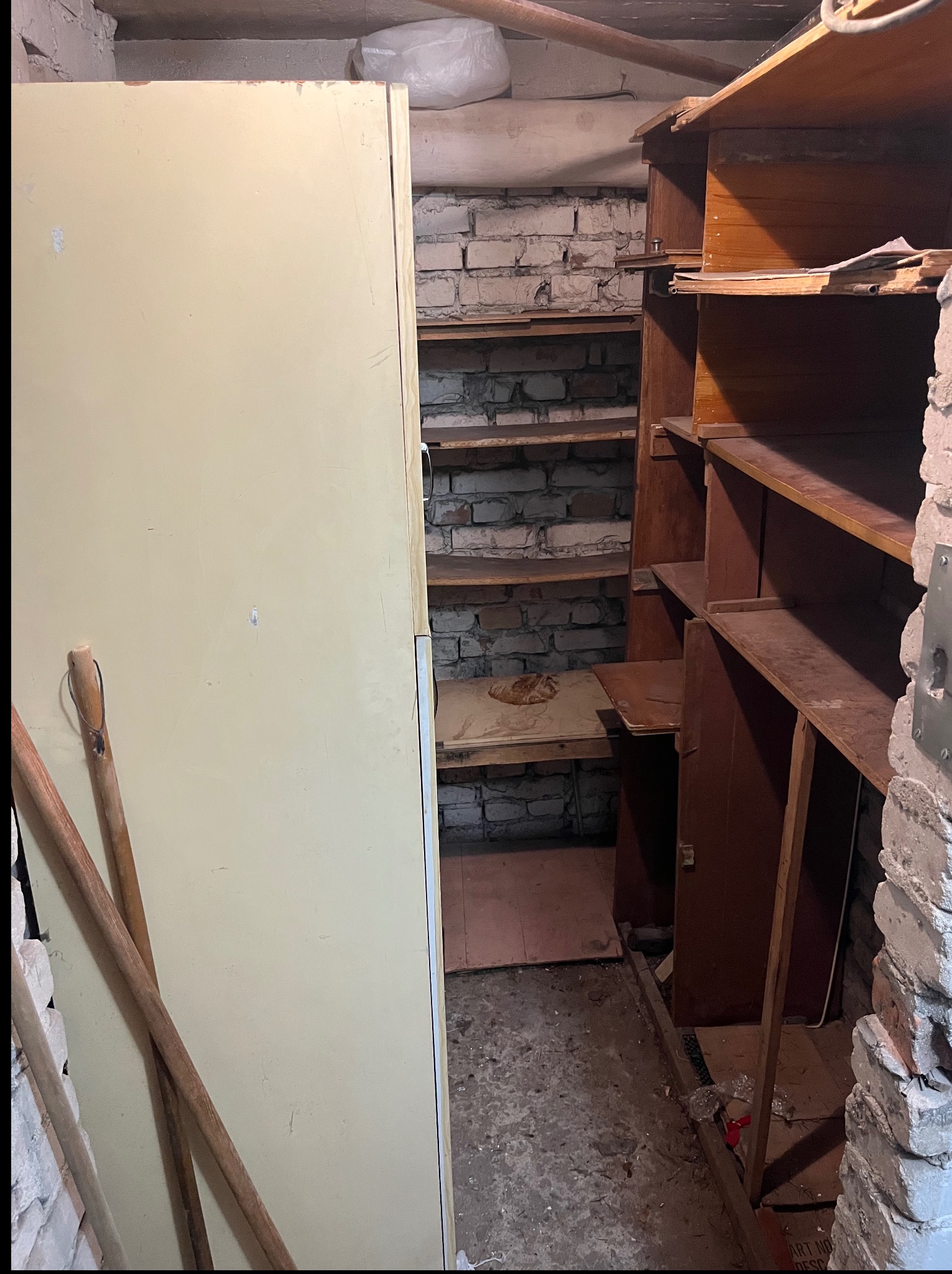 Zdemontowane, drewniane regały w piwnicy z ceglanymi ścianami. Obok stoi lodówka i leżą drewniane kije. Widoczny gruz i resztki po demontażu.