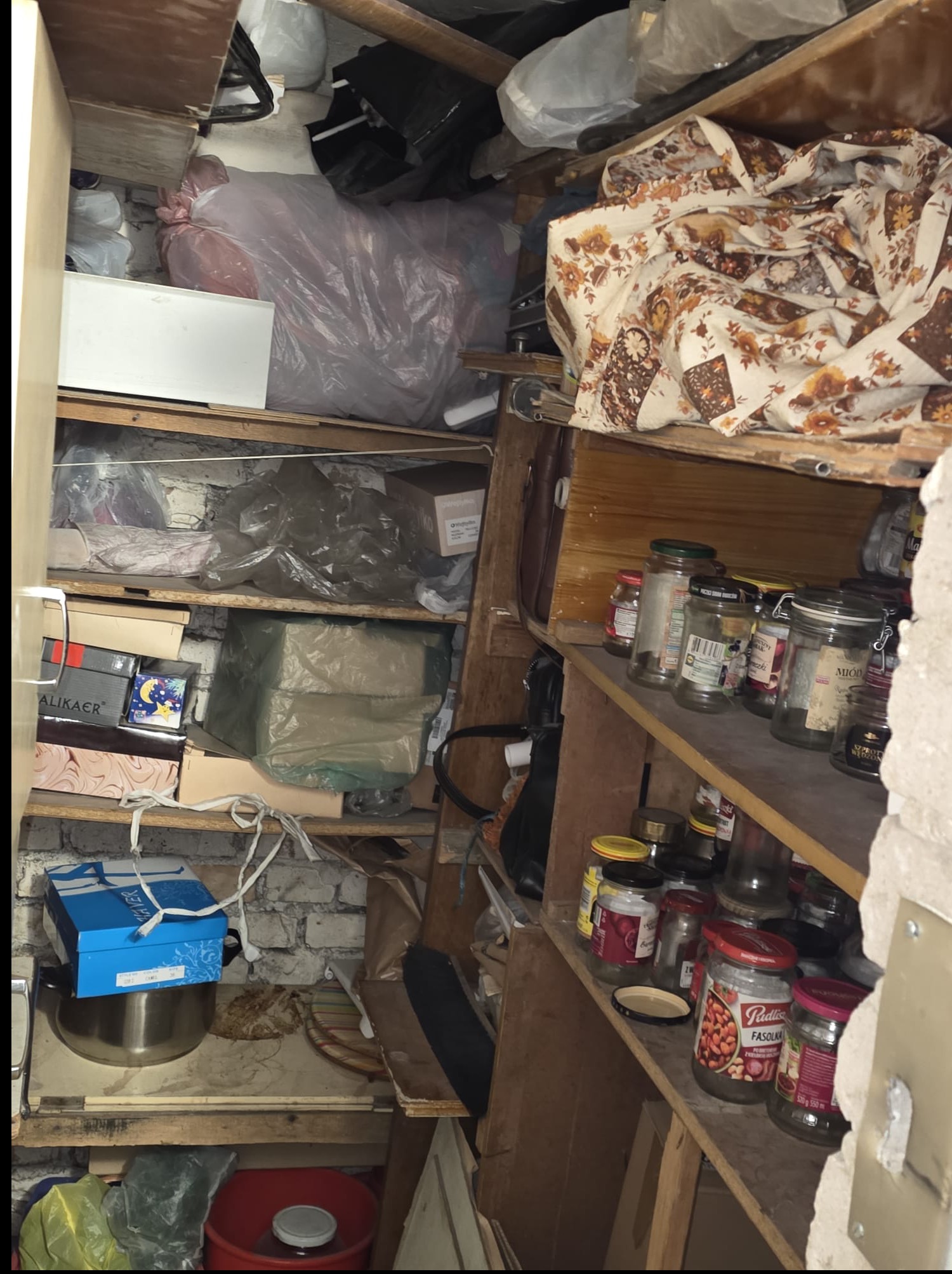 Zastawiona półka w pomieszczeniu z różnymi przedmiotami, w tym słoikami, pudełkami i workami. Widoczne elementy muru i drewniane półki. Nieporządek.