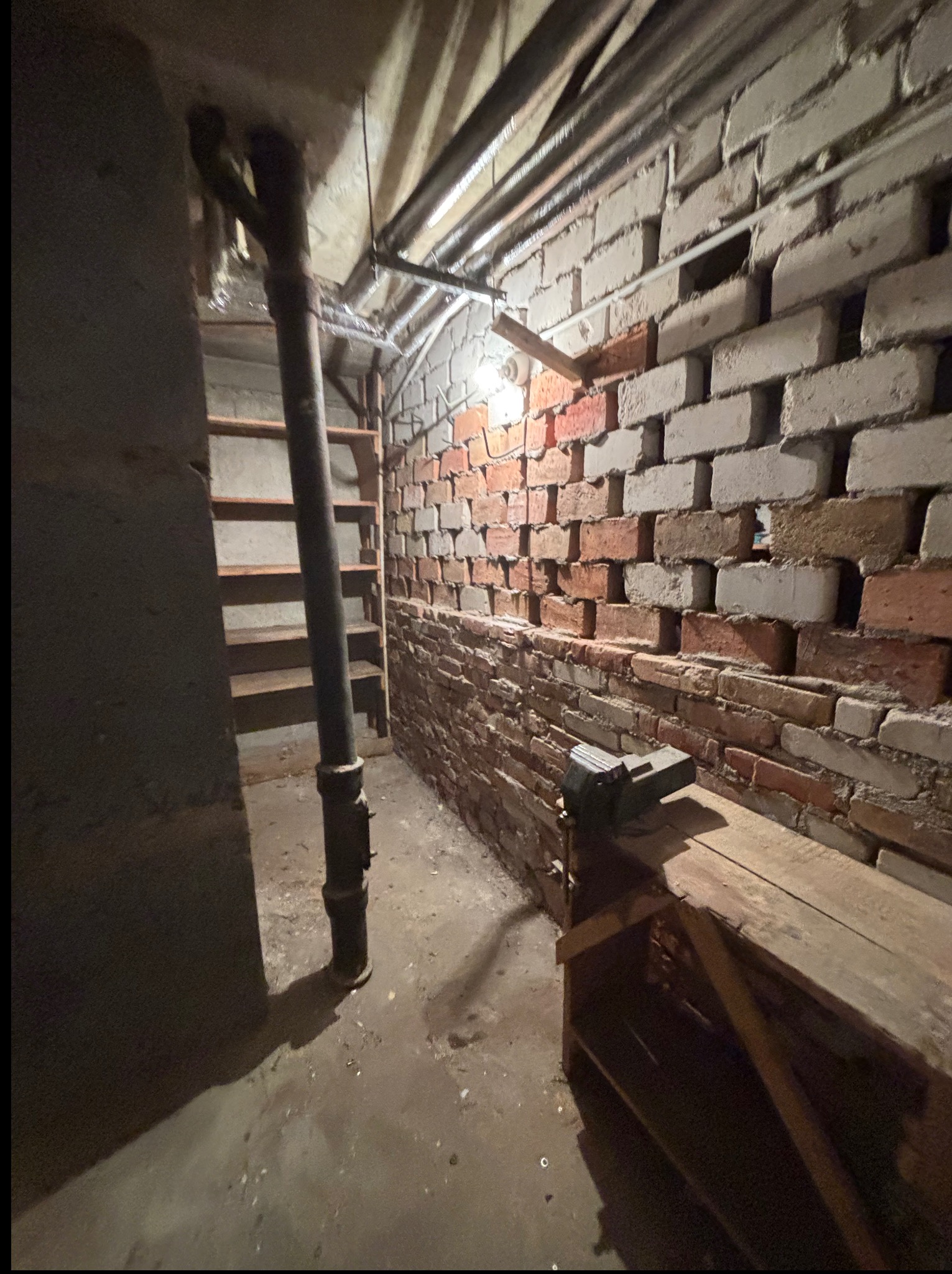 Surowe wnętrze piwnicy z ceglaną ścianą, drewnianym stołem warsztatowym i metalową rurą. Widoczne schody i surowe oświetlenie. Klimat starego warsztatu.