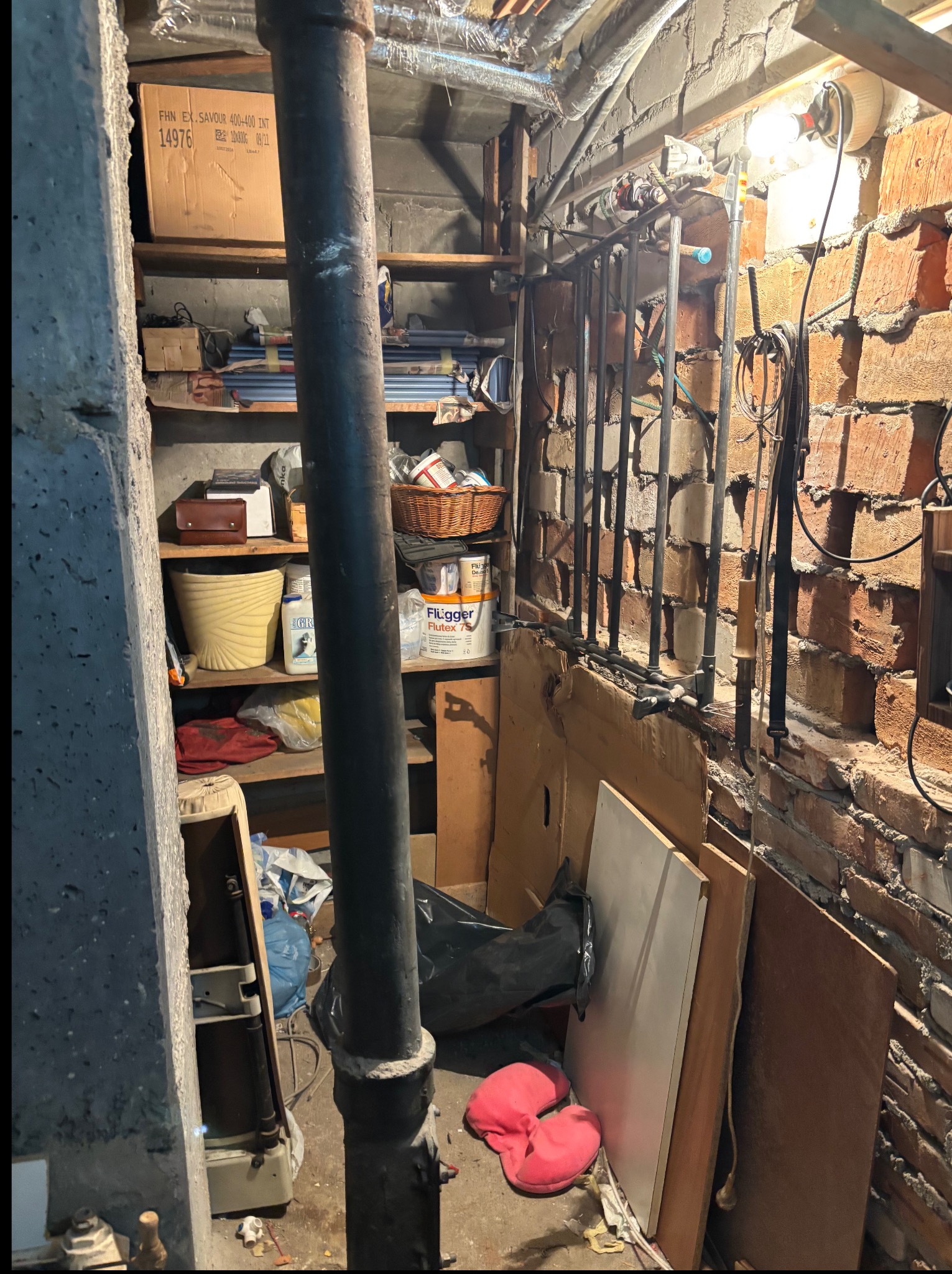 Zagracone pomieszczenie z półkami, rurami i ceglaną ścianą. Widoczne kartony, wiadra, narzędzia i inne przedmioty. Panuje ogólny nieporządek, typowy dla piwnicy lub strychu.