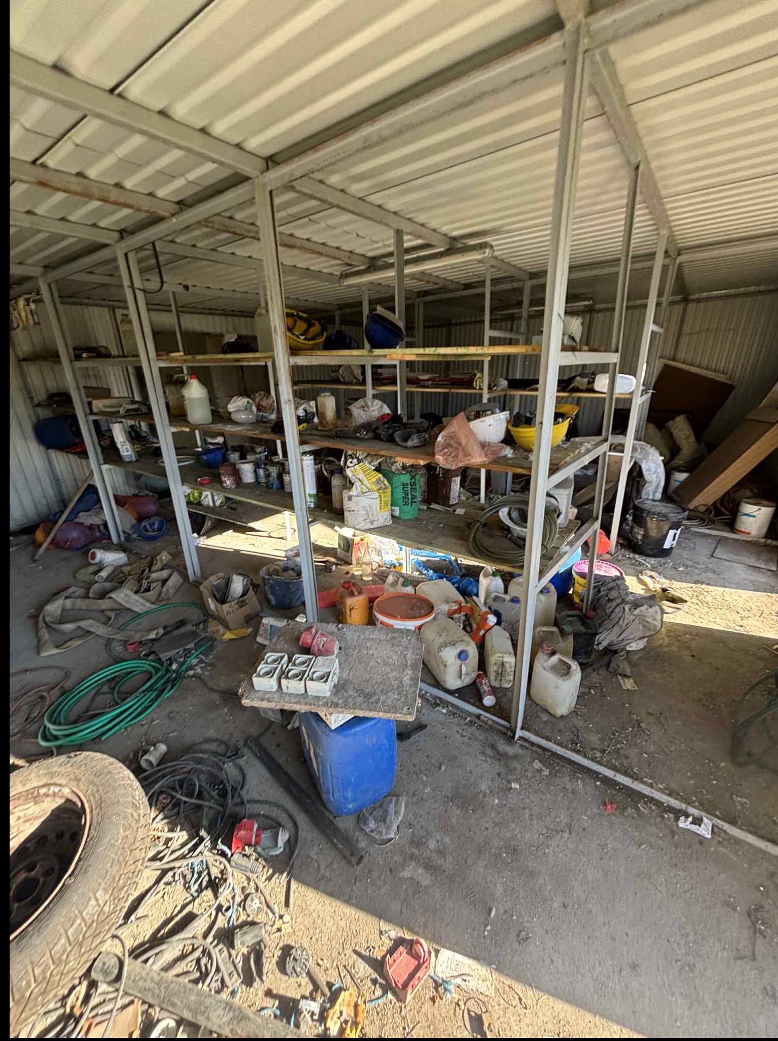 Zabałaganiona wiata z metalową konstrukcją i półkami, na których leżą różnorodne przedmioty, w tym pojemniki, wiadra i narzędzia. Na podłodze opona i kable.