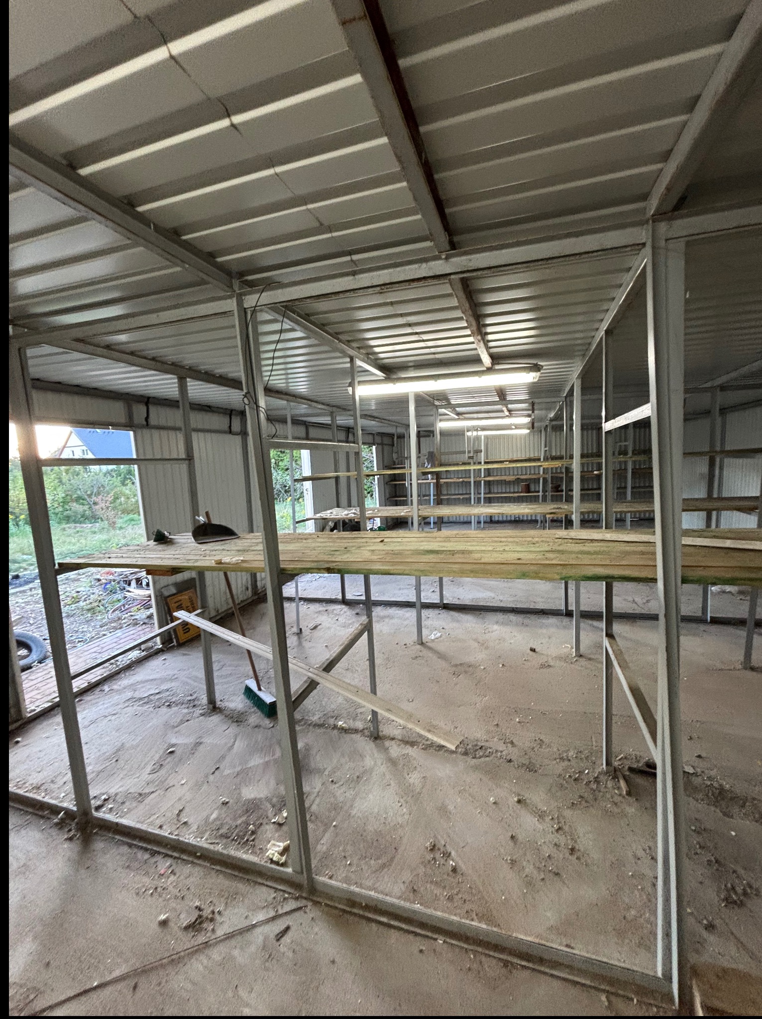 Wnętrze hali w trakcie opróżniania: widoczne metalowe konstrukcje, drewniane deski, resztki po remoncie i narzędzia. Oświetlenie sufitowe, widok na zewnątrz przez otwarte drzwi.