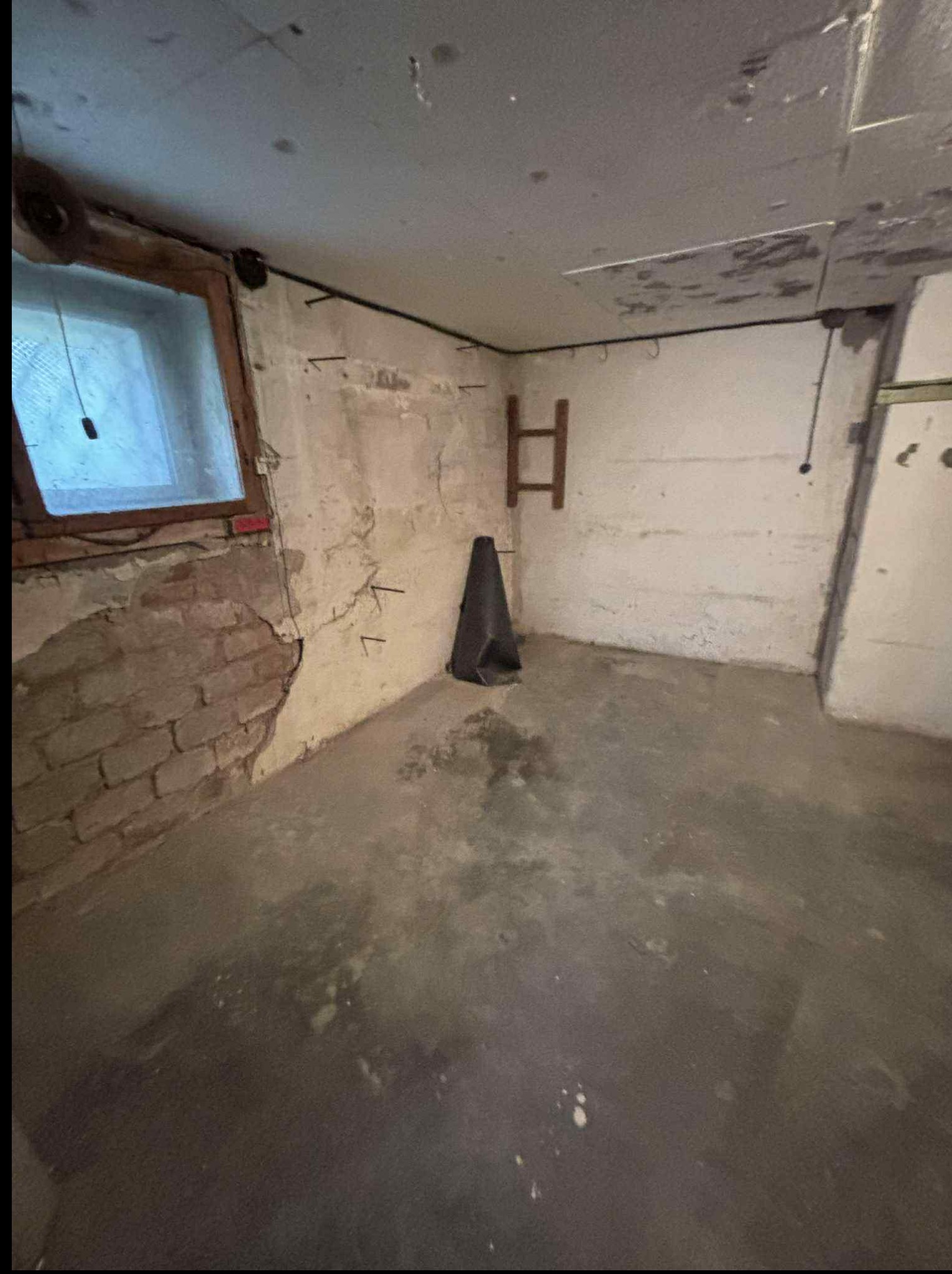 Piwnica z oknem, fragmentem odsłoniętej cegły i betonową podłogą. Na ścianie widoczne ślady po instalacjach i hak. W rogu stoi czarny stożek.