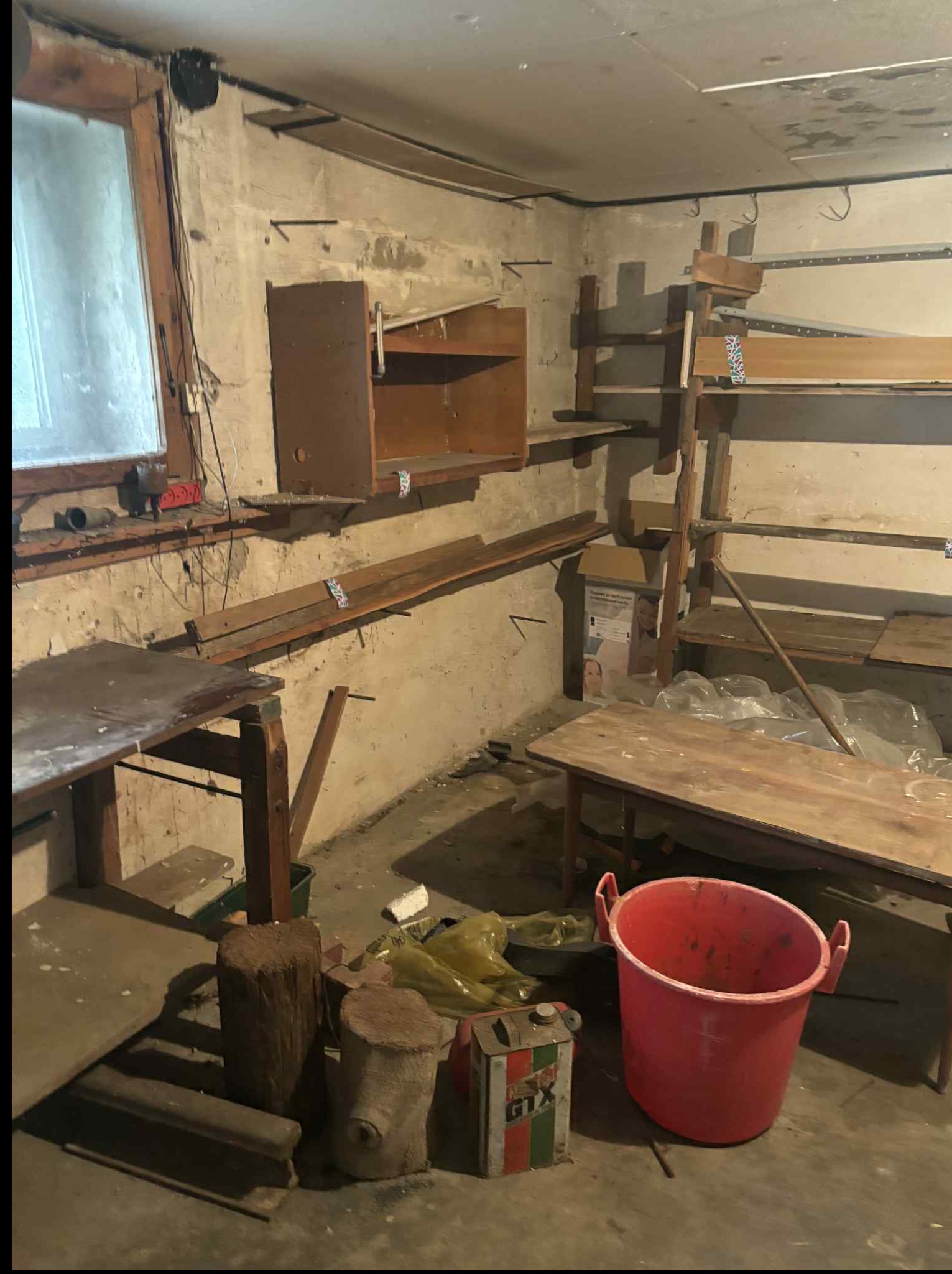 Zagracone pomieszczenie z drewnianymi półkami, stołami i wiadrem. Widoczne ślady wilgoci na ścianach i resztki przedmiotów. Nierównomierne oświetlenie.
