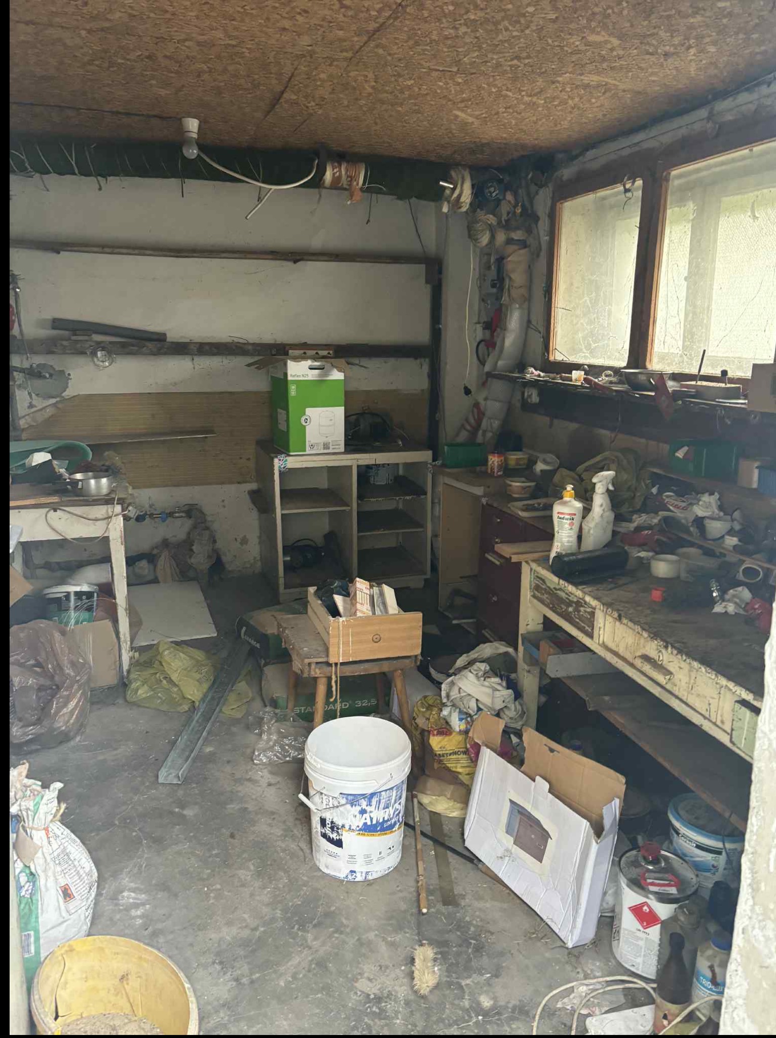 Zagracony warsztat lub pomieszczenie gospodarcze z oknem, widoczne narzędzia, puszki z farbą, kartony i inne przedmioty. Panuje ogólny nieporządek.
