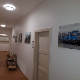 2P Usługi wykończeniowo budowlane - Korytarz z białymi ścianami, na których wiszą zdjęcia. Na podłodze leżą kartony. Na ścianie wisi zdjęcie lokomotywy. Widoczne drzwi i oświetlenie sufitowe.