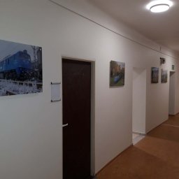 2P Usługi wykończeniowo budowlane - Korytarz biurowy z białymi ścianami, na których wiszą obrazy. Widoczne drzwi i kartony na podłodze. Oświetlenie sufitowe.