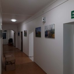 2P Usługi wykończeniowo budowlane - Korytarz biurowy z białymi ścianami, na których wiszą obrazy. Widoczny znak 'Wyjście Ewakuacyjne' nad drzwiami i karton na podłodze. Ujęcie z perspektywy.