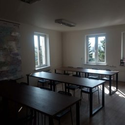 2P Usługi wykończeniowo budowlane - Jasne wnętrze sali lekcyjnej z drewnianymi stołami i krzesłami, mapa na ścianie, trzy okna z widokiem na zieleń. Parkiet na podłodze, oświetlenie sufitowe.