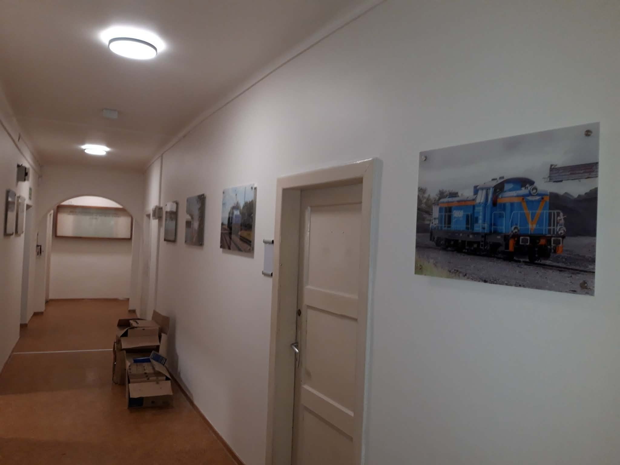 Korytarz z białymi ścianami, na których wiszą zdjęcia. Na podłodze leżą kartony. Na ścianie wisi zdjęcie lokomotywy. Widoczne drzwi i oświetlenie sufitowe.