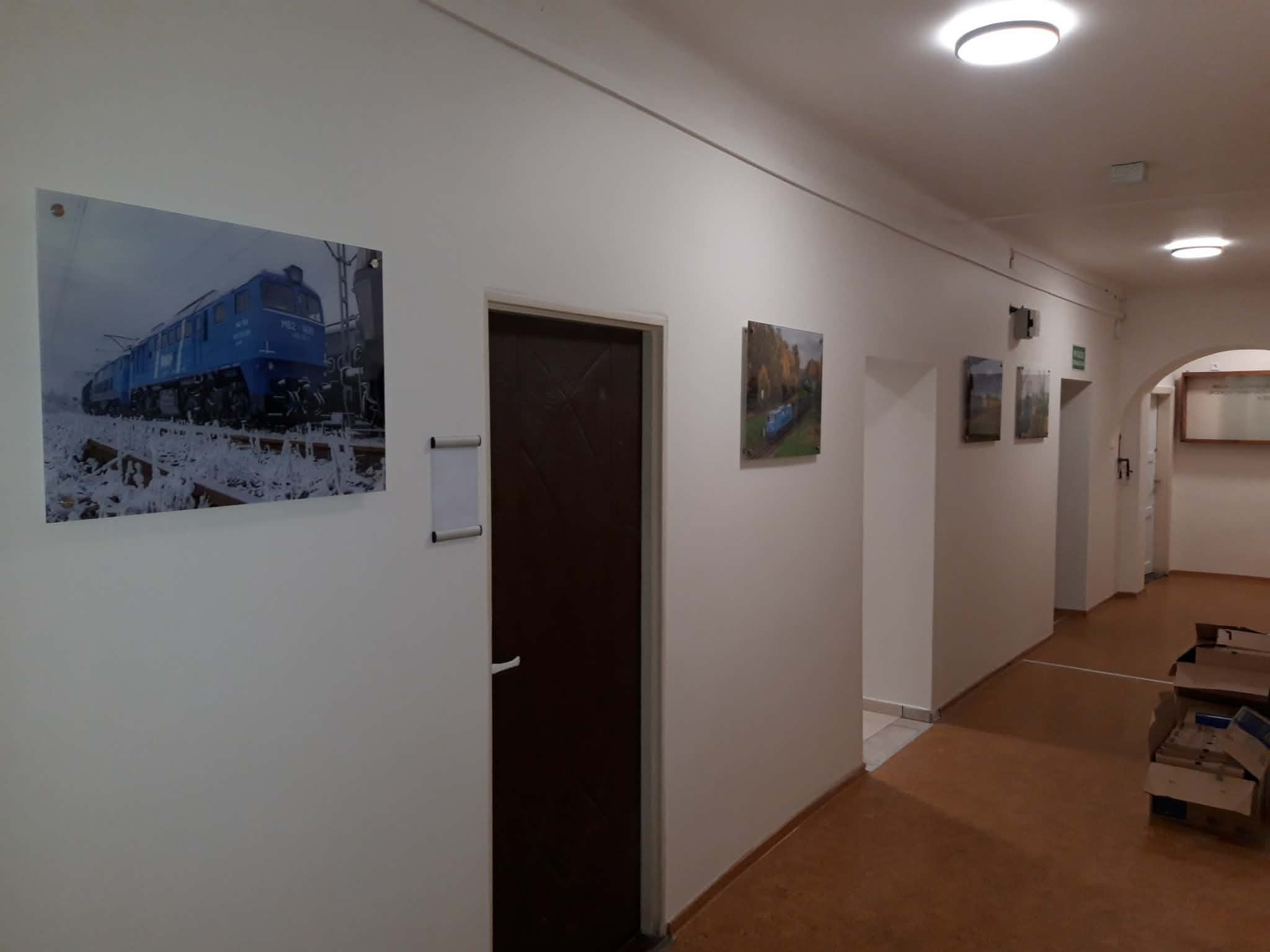 Korytarz biurowy z białymi ścianami, na których wiszą obrazy. Widoczne drzwi i kartony na podłodze. Oświetlenie sufitowe.