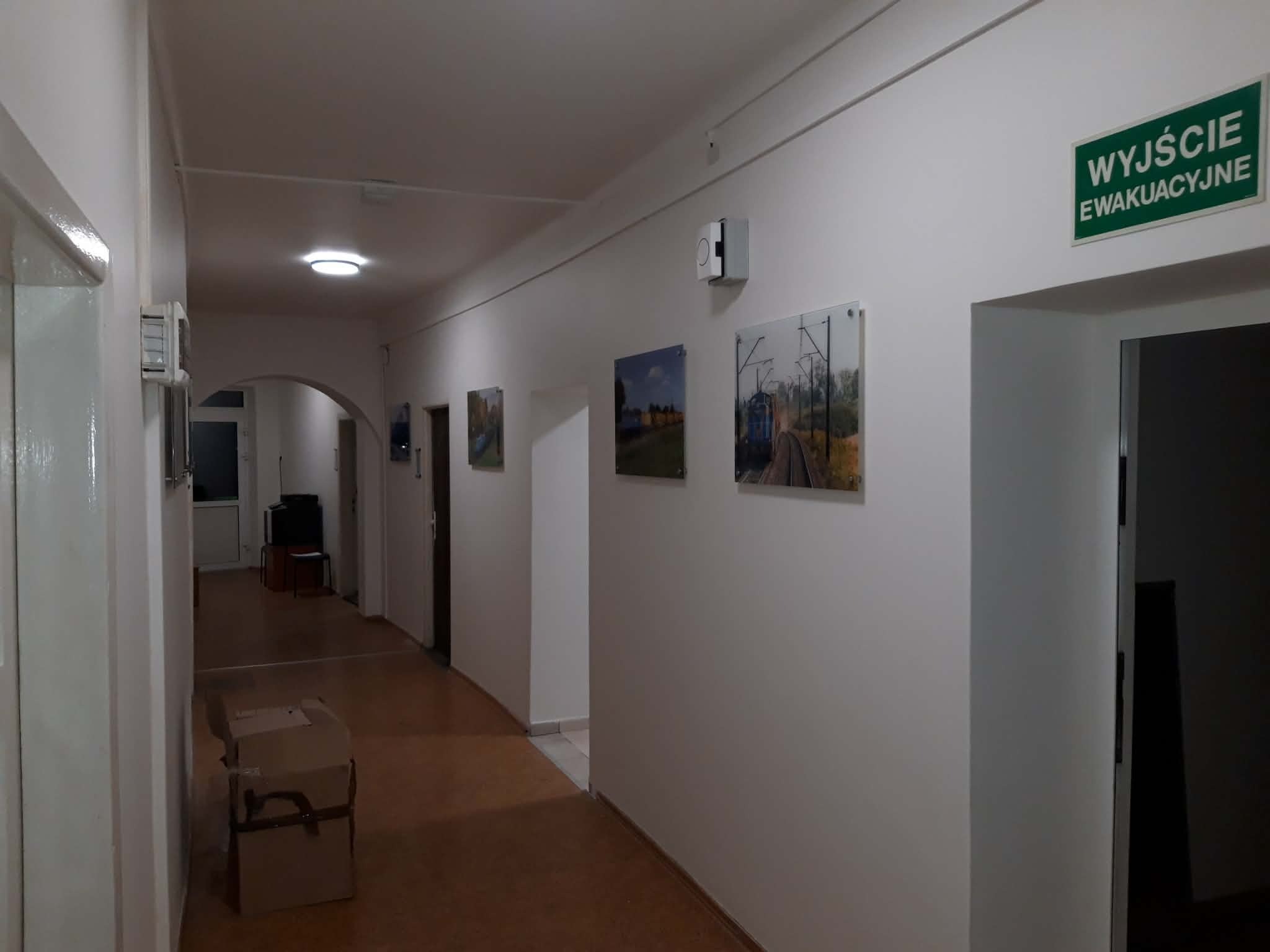 Korytarz biurowy z białymi ścianami, na których wiszą obrazy. Widoczny znak 'Wyjście Ewakuacyjne' nad drzwiami i karton na podłodze. Ujęcie z perspektywy.