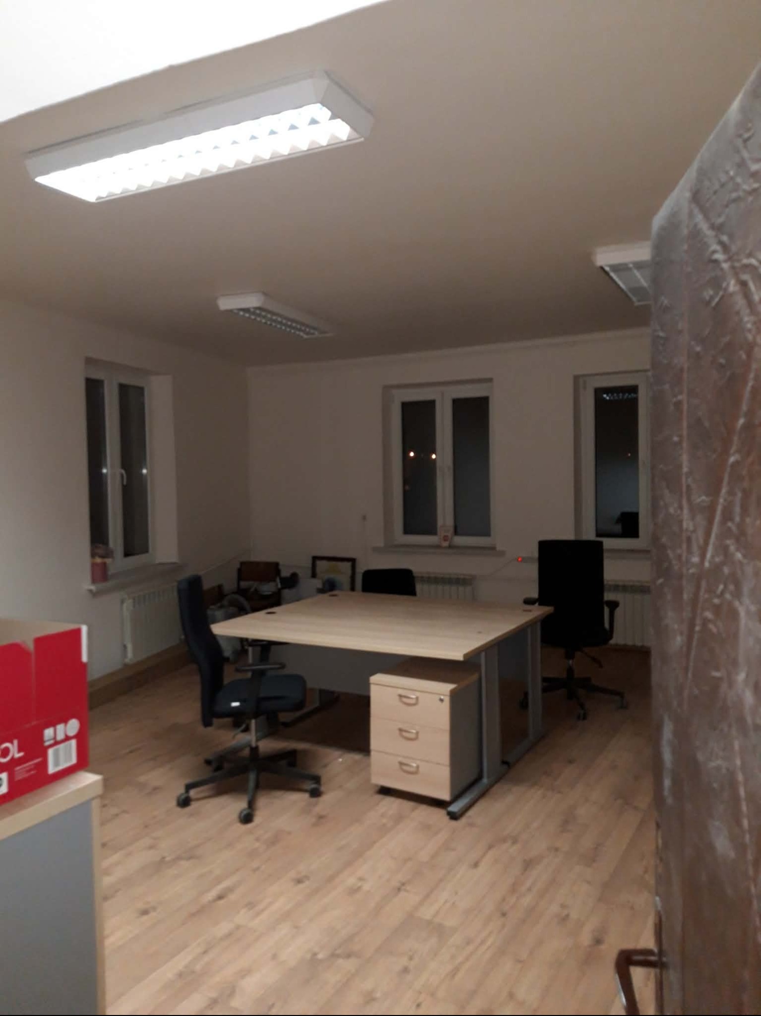 Jasne biuro z drewnianą podłogą, biurkami i krzesłami. Widoczne okna z białymi ramami i oświetlenie sufitowe. Częściowo widoczna ściana z dekoracyjną tapetą.