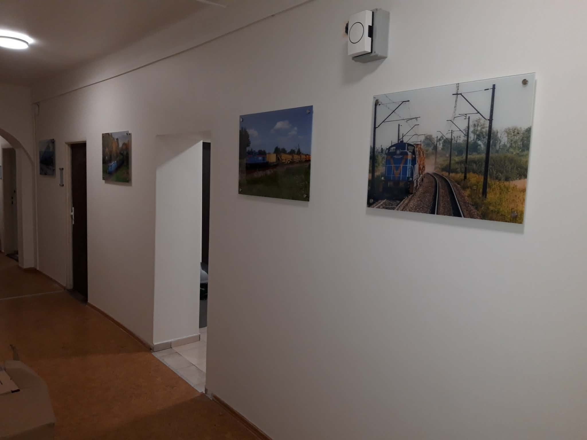Korytarz biura w Dębicy z obrazami pociągów na szklanych panelach, zamontowanych na białej ścianie. Widoczne drzwi i fragment podłogi. Oświetlenie sufitowe.
