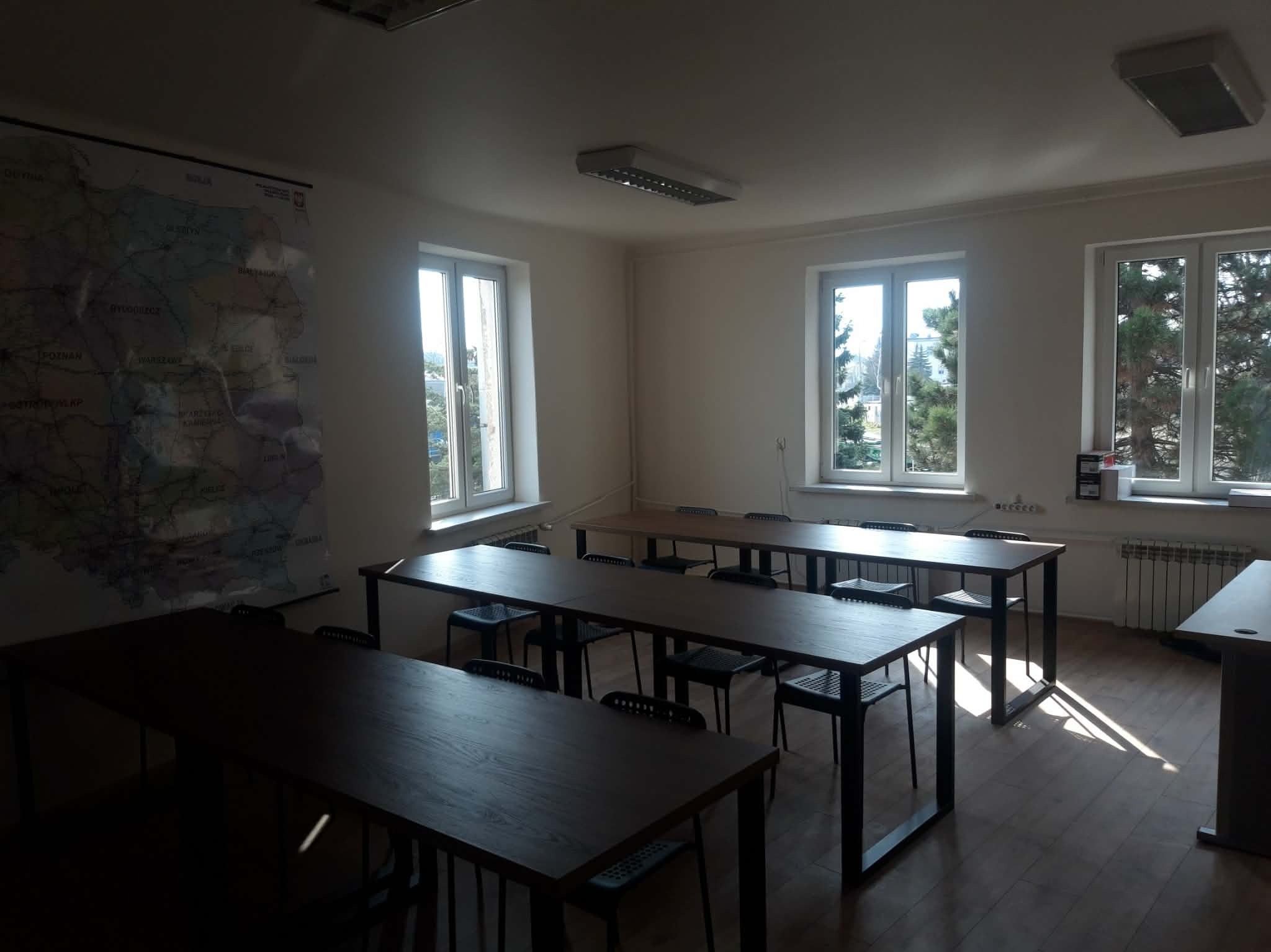 Jasne wnętrze sali lekcyjnej z drewnianymi stołami i krzesłami, mapa na ścianie, trzy okna z widokiem na zieleń. Parkiet na podłodze, oświetlenie sufitowe.