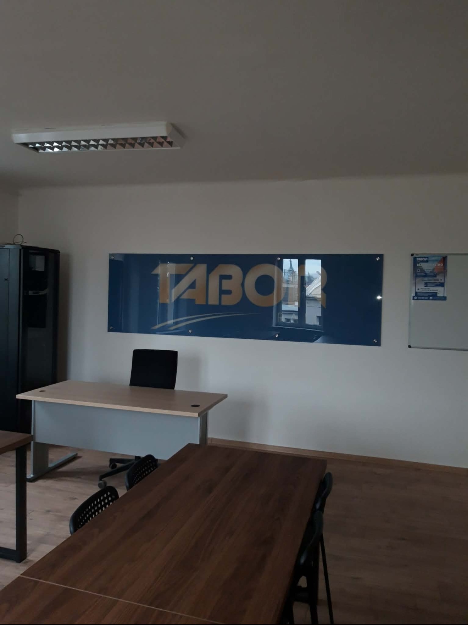 Biuro z drewnianą podłogą, biurkami i krzesłami. Na ścianie logo firmy TABOR na niebieskim tle, obok tablica suchościeralna. Widoczna lampa sufitowa.