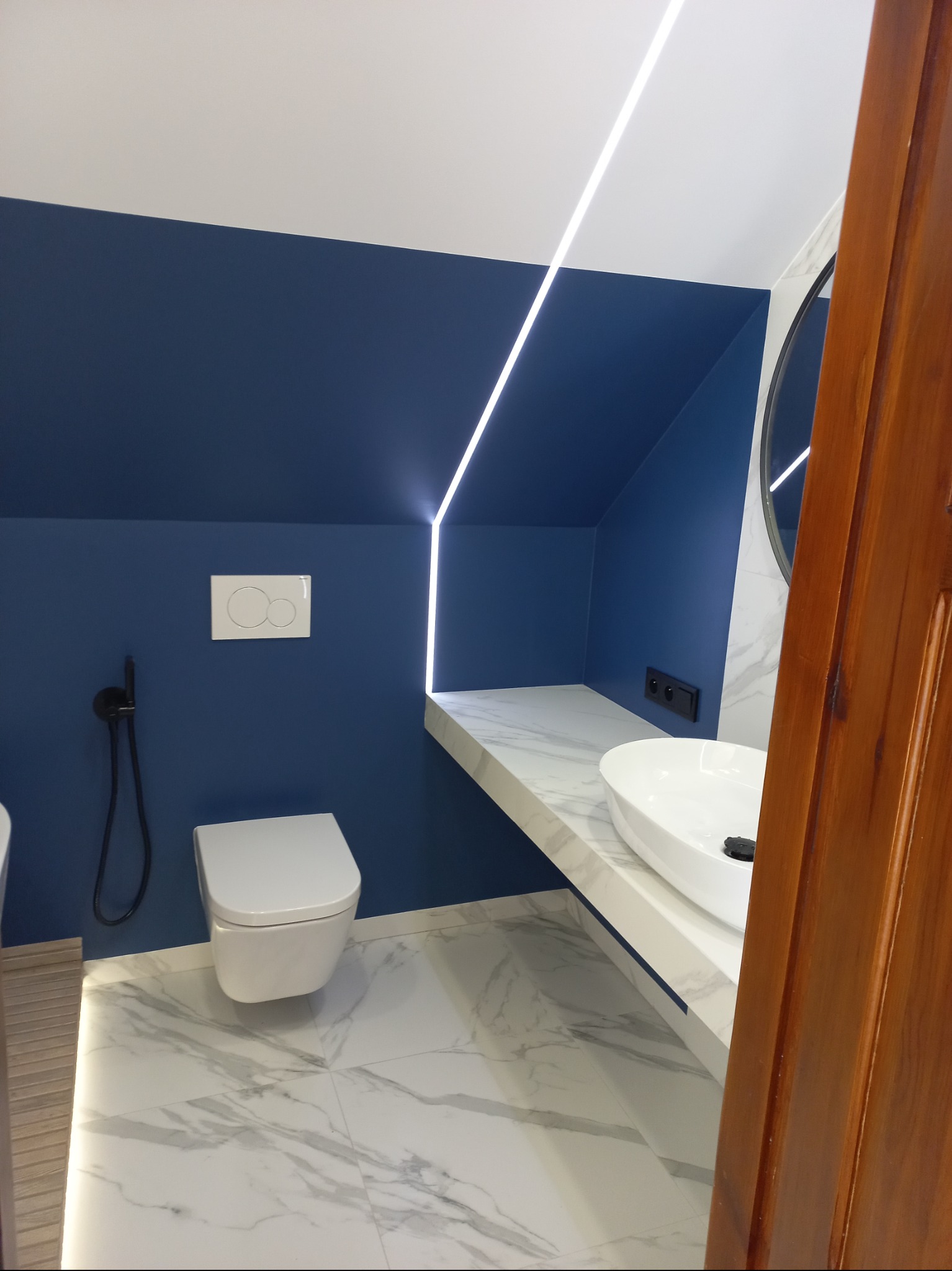 Niebieska łazienka na poddaszu z marmurową posadzką i blatem, oświetleniem LED wzdłuż ściany i nowoczesną armaturą. Umywalka owalna, toaleta podwieszana.