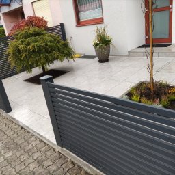 Iron Tech Solutions - Nowoczesne, poziome ogrodzenie panelowe w kolorze grafitowym, oddzielające posesję od chodnika. Widoczne płytki chodnikowe i roślinność w ogrodzie. Ujęcie z niskiej perspektywy.