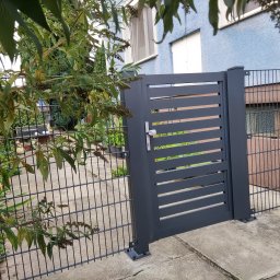 Iron Tech Solutions - Nowoczesna, grafitowa furtka z poziomymi szczeblami, wkomponowana w ogrodzenie panelowe. Widoczne detale montażu i zamek. W tle zieleń ogrodu i elewacja budynku.