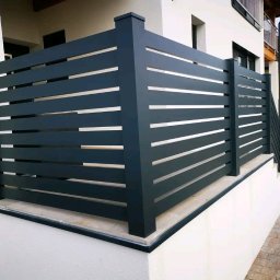 Iron Tech Solutions - Nowoczesne, poziome ogrodzenie metalowe w antracytowym kolorze, z subtelnymi prześwitami, zamontowane na niskim murku przy schodach zewnętrznych budynku.