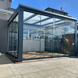 Iron Tech Solutions - Szklana zabudowa balkonu z przesuwnymi drzwiami, widok z zewnątrz. Nowoczesna konstrukcja z ciemną ramą i drewnianą podłogą. Jasne tło nieba i budynków.