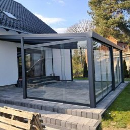 Iron Tech Solutions - Szklana zabudowa tarasu z szarą ramą, widok z zewnątrz. Nowoczesna konstrukcja z przesuwnymi panelami szklanymi na kamiennej posadzce, zieleń w tle.