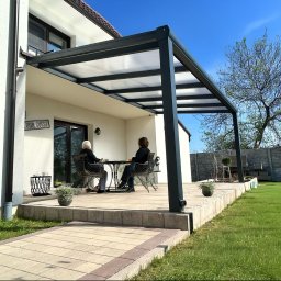 Iron Tech Solutions - Nowoczesna, grafitowa pergola metalowa z przezroczystym zadaszeniem, przylegająca do białego domu. Dwie osoby siedzą przy stoliku na tarasie. Widok na ogród i błękitne niebo.