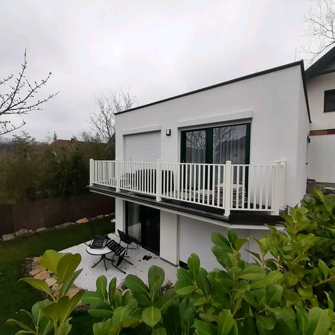 Biała balustrada balkonowa z pionowymi szczeblami, nowoczesny budynek z roletami, widok z dołu, zieleń w tle i na pierwszym planie, pochmurne niebo.