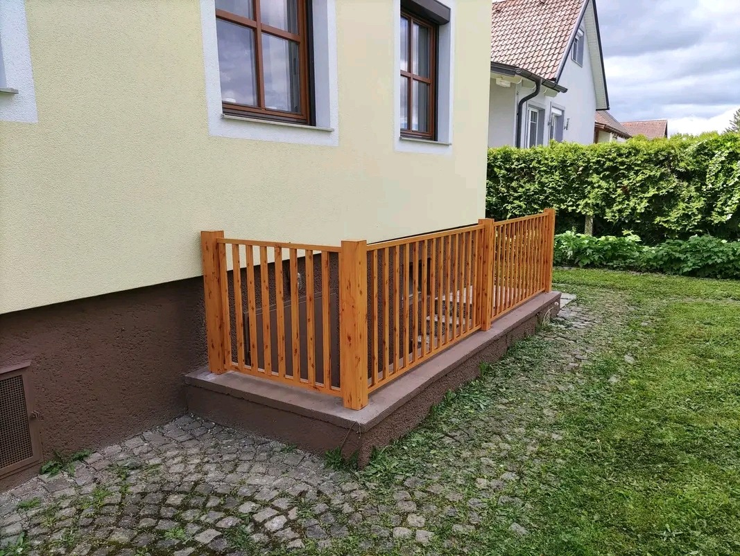 Drewniana balustrada zabezpieczająca niskie okno piwniczne w domu jednorodzinnym. Widok z zewnątrz, trawnik i kostka brukowa.