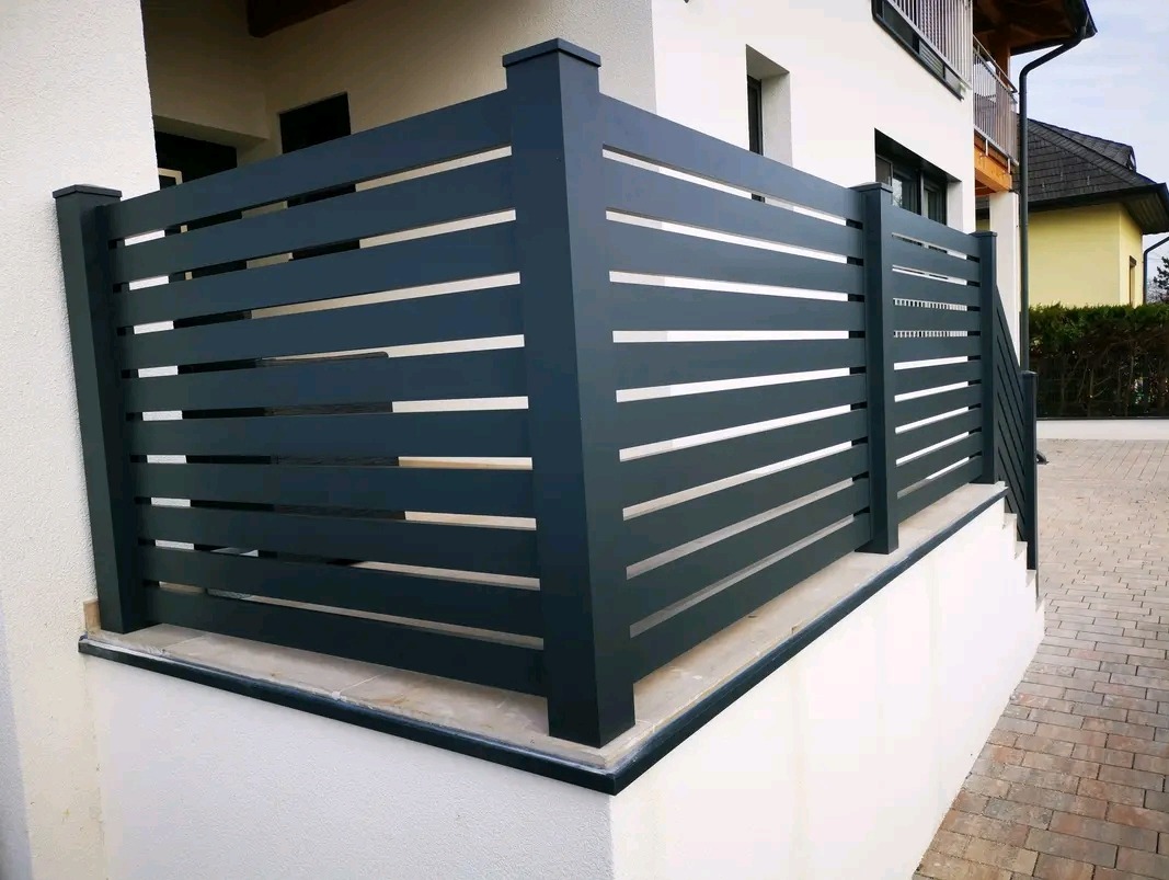 Nowoczesne, poziome ogrodzenie metalowe w antracytowym kolorze, z subtelnymi prześwitami, zamontowane na niskim murku przy schodach zewnętrznych budynku.