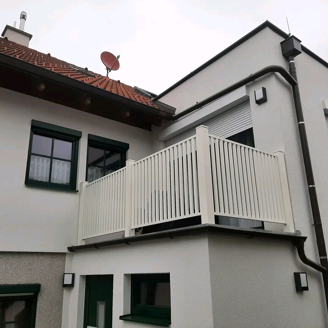 Biała balustrada balkonowa z pionowymi szczeblami, zamontowana na narożnym balkonie budynku z jasną elewacją i ciemną stolarką okienną. Widok z dołu.